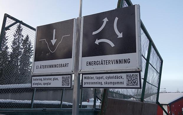 Två svarta skyltar på en stolpe. Den ena är med en vit pil som pekar ner i en grop, under står det "ej återvinningsbart", den andra är det tre böjda pilar som pekar från varandra och under står det "energiåtervinning"