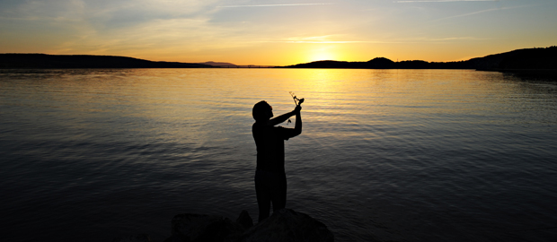En fiskare står i motljus och fiskar. Solnedgång i bakgrunden.
