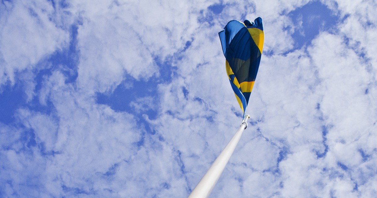 Svenska flaggan på flaggstång mot blå himlen med vita stråk av moln