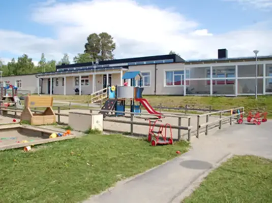 en grå byggnad i bakgrunden, i förgrunden lekytor utomhus med lekställningar och en stor sandlåda