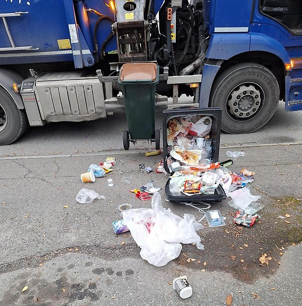Blå sopbil lyfter ett kärl med brunt lock. Bredvid ligger ett vält sopkärl där plastförpackningarna har vällt ut.