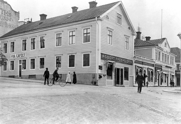 Gammal, svartvit bild på ett ljust trähus i två våningar med liggande panel och tre skorstenar.