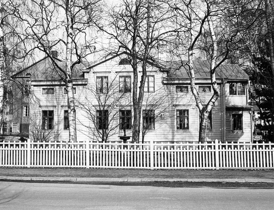 Gammal, svartvit foto på en villa i två våningar, ljus liggande panel och mörka fönster.