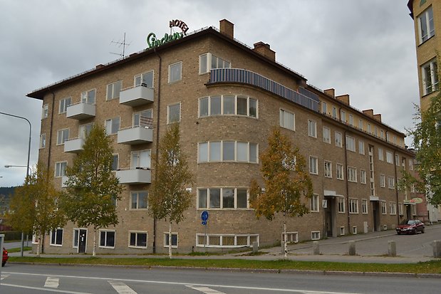 Byggnad i fem våningar i gult tegel. Rundat hörn och balkonger i vit plåt.