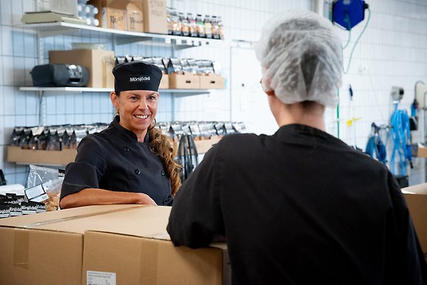 Anna står och lutar sig mot kartonger och pratar med person med ryggen vänd mot kameran