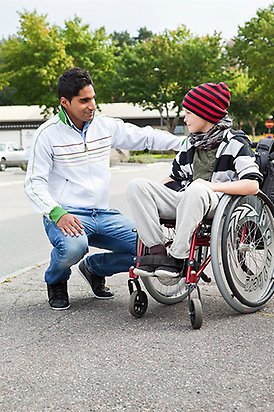 En vuxen man som sitter på huk ler mot ett barn i en rullstol.