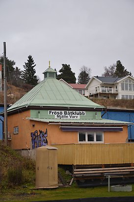 Frösö 2:40. Byggnad i orange puts med grönt plåttak, bruna dubbeldörrar, utbyggnad med sluttande tak och tre små fönster. Står Frösö Båtklubb på fasaden.