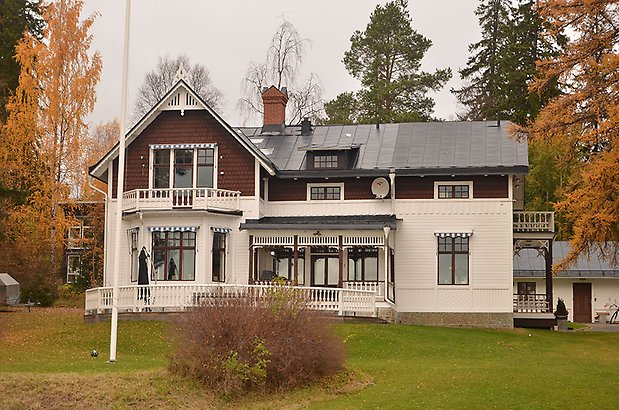 Solskiftet 7. Villa med vit liggande panel på undervåningen, och brun fiskfjällspanel på övervåningen. Bruna fönster med spröjs och svart plåttak. 