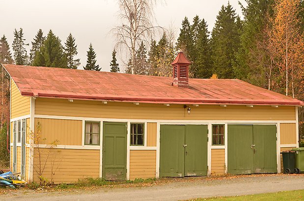 Ägostyckningen 7. Byggnad i gul, liggande panel, dörrar och garageportar i grönt och gröna fönster med spröjs. Rött plåttak.