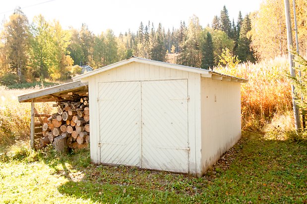 Byggnad i vit panel med dubbelport, med snedtak till vänster med ved under.