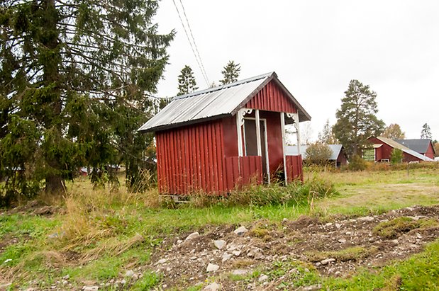 Liten byggnad i röd panel med förstukvist med snickarglädje och svart dörr.