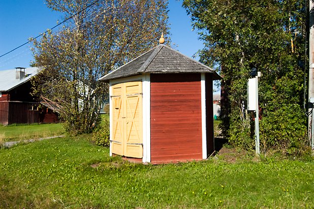 Litet åttakantigt hus med röd panel och gul dubbelport.