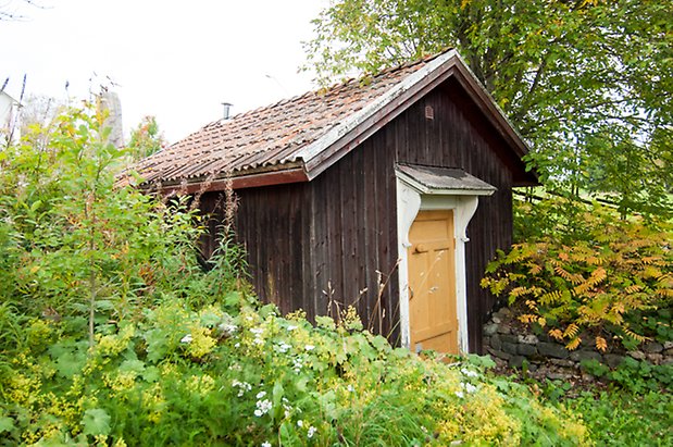 Boda 1:15. Liten byggnad i grånad panel med gul dörr.