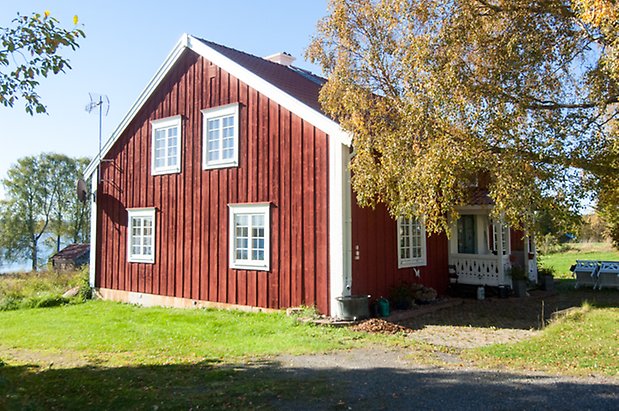 Handog 2:34. Hus i röd, stående panel. Med vit förstukvist med snickarglädje och fönster med vita spröjs.