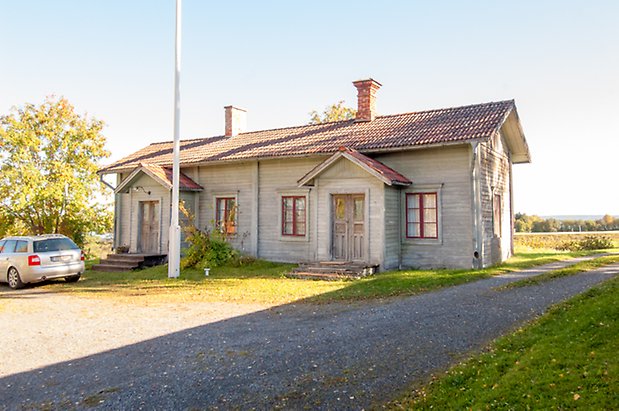 Hökbäck 2:8. Hus i liggande grå panel med två grå dubbeldörrar och fönster med röda spröjs och två skorstenar.