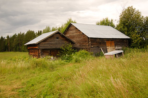 Hara 7:29. Timrat, brunt härbre med timrad, brun byggnad bredvid med ljusbrun dubbelport.