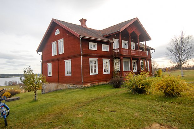 Oldberg 1:5. Timrad, röd byggnad i tre våningar med röd förstukvist med balkong ovanför med snickarglädje, fönster med vita spröjs.