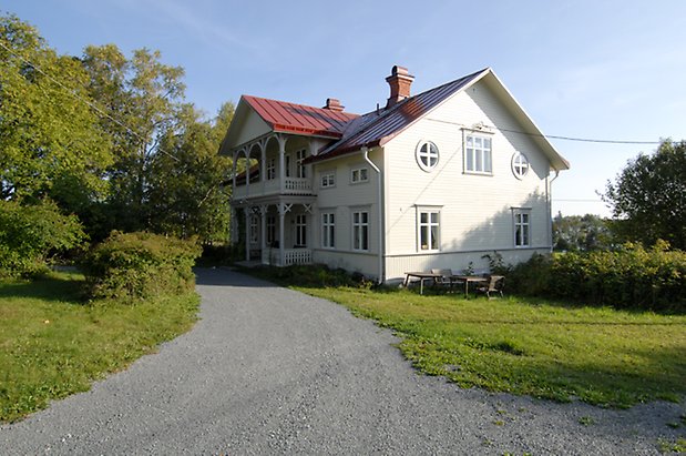 Jälsta 1:2. Villa i två våningar i vit, liggande panel med grå detaljer, förstukvist med balkong ovanför med snickarglädje.