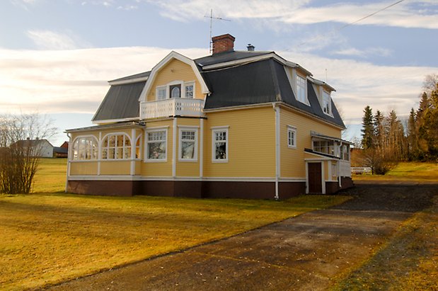 Fugelsta 2:26. Villa i två våningar i gul, liggande panel, punschveranda, balkong med snickarglädje och fönster med vita spröjs.