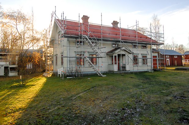 Marieby-Bye 2:18. Villa i två våningar i vit, liggande panel med grå och mörka detaljer, förstukvist. Huset har byggnadsställning runt sig.