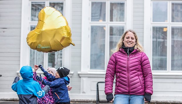 En tjej med täckjacka står och tittar mot kameran. Lekande barn i bakgrunden