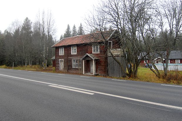 Kläppe 3:8. Hus i brunt timmer i två våningar, fönster med vita spröjs, förstukvist med vita pelare, balkong med vit snickarglädje på kortsidan. Huset ligger precis vid en väg.