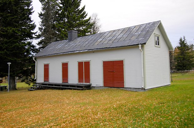 Häggenås Prästbord 1:7. Byggnad i vit, liggande panel med två röda dubbelportar, och två portar, skorsten.