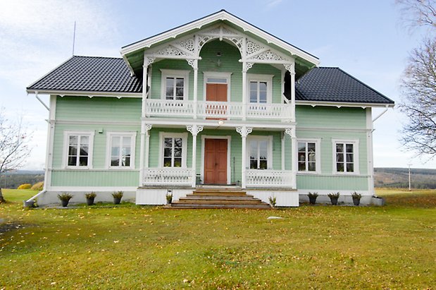 Österåsen 1:32. Villa i ljusgrön, liggande panel med vita detaljer, förstukvist med balkong ovanför med vit snickarglädje, två dubbeldörrar i brunt, fönster med vita spröjs.