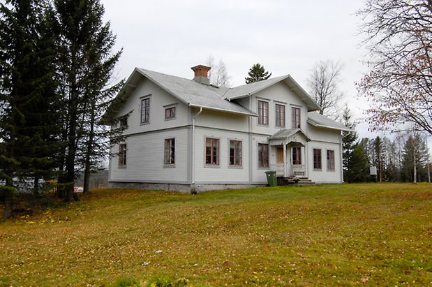 Kogsta 2:27. Villa i vit, liggande panel med grå detaljer, fönster med röda spröjs, takkupa med tre fönster. Förstukvist med snickarglädje och gul dörr.