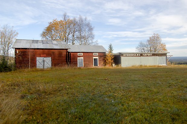 Storhögen 4:15. En röd, timrad byggnad med blå garageport, en timrad, röd byggnad med två blå portar, byggnad med metallportar och texten "Storhogens Airport".