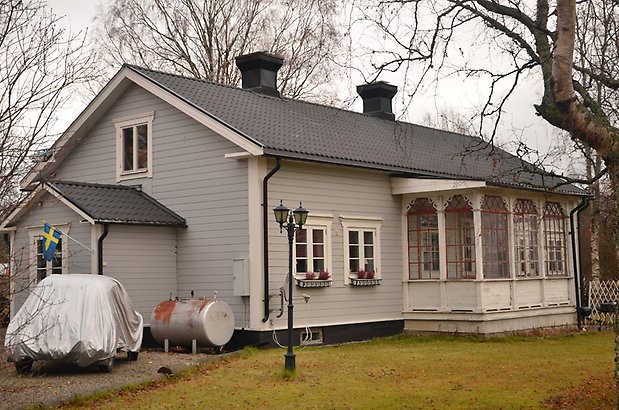 Draget 6. Bostadshus med ljusgrå liggande panel, vit punschveranda, vita fönster med röda spröjs.