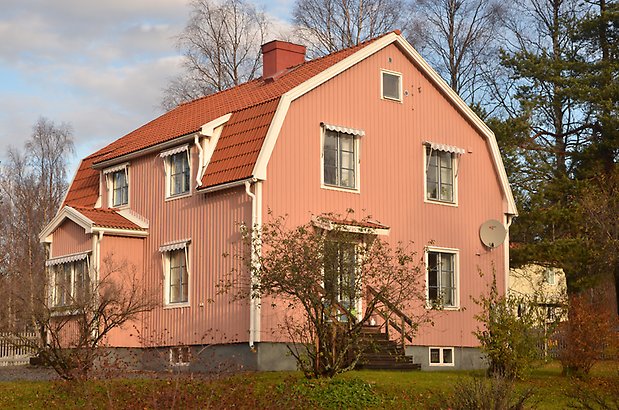 Ask 6. Villa rosa träpanel med vita fönster och blå spröjs och ett burspråk.