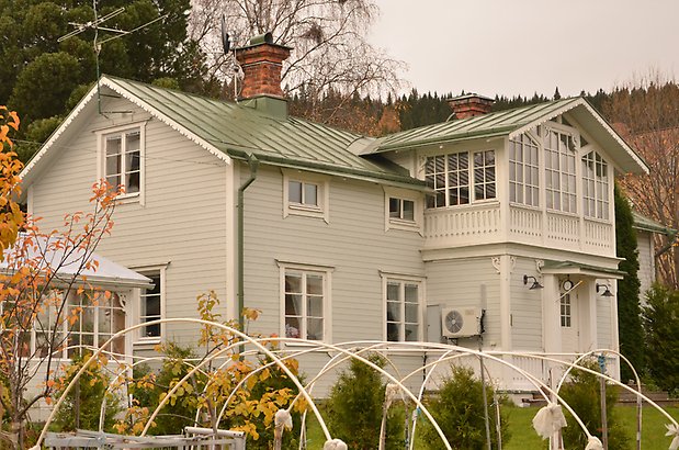 Muraren 7. Villa i grå liggande panel och vita fönster med spröjs, inglasad balkong med snickarglädje.