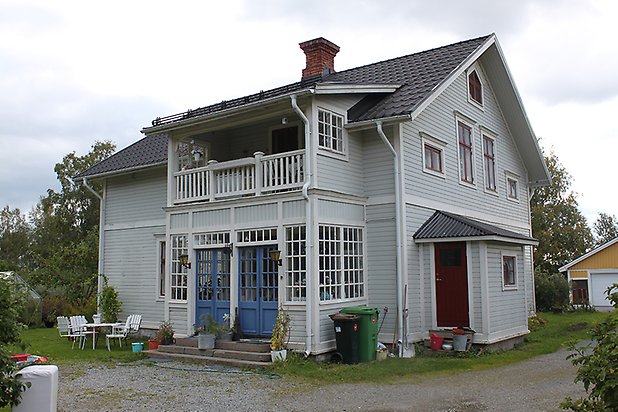 Brunflo-Änge 1:24. Villa i ljusgrå, liggande panel med vita fönster med röda spröjs. Punschveranda med blå dörrar.