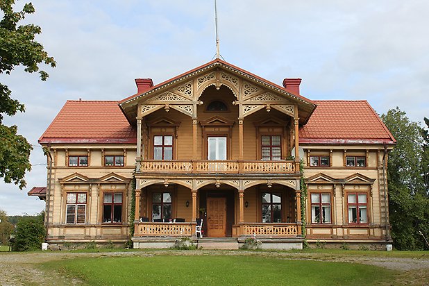 Brunflo-Änge 1:118. Villa i ljusgul liggande panel med träfärgade detaljer och balkong och veranda med snickarglädje.