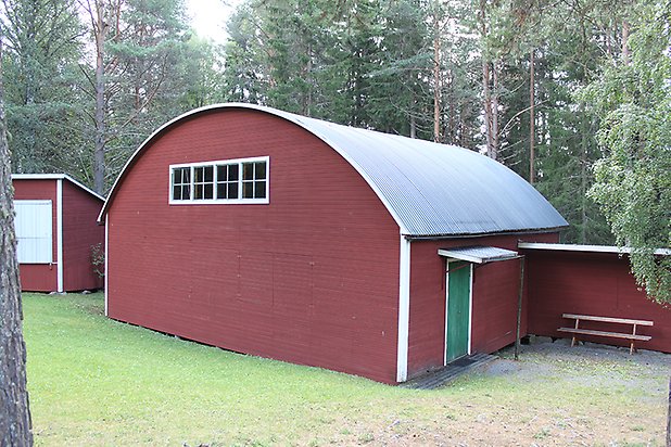 Häggesta 1:63. Byggnad i röd, liggande panel med grön dörr och små vita fönster med gröna spröjs, svart, välvt plåttak.