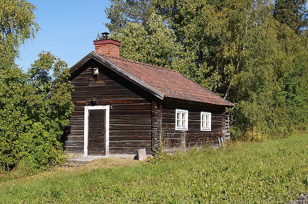 Torvalla 4:50. Stuga i svart timmer med fönster med vita spröjs, svart dörr, skorsten i tegel.