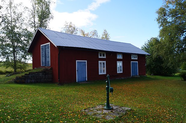 Lunne 3:13. Ladugård i rött timmer och panel och tre blå dubbelportar, fönster med vita spröjs.