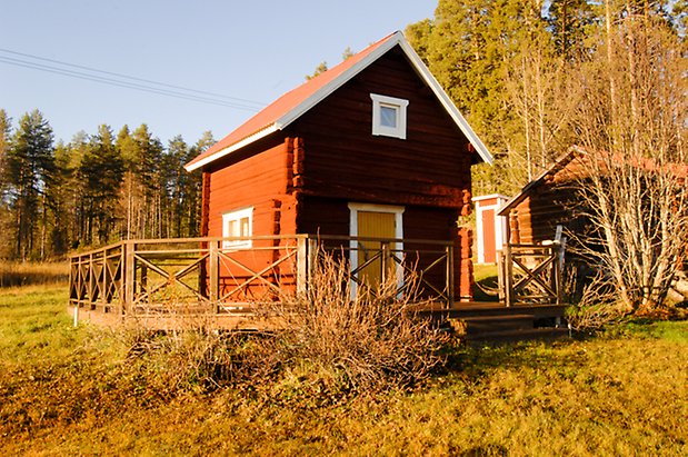 Lockne-Berge 1:7. Rött, timrat härbre med gul dörr och vita fönster. Ett trädäck med staket runt härbret.