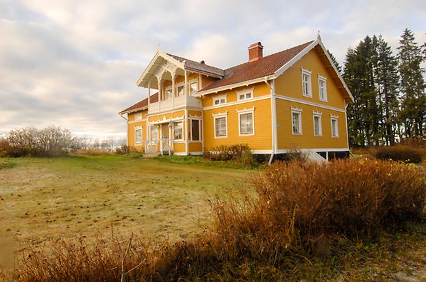 Lockne-Änge 2:9. Villa i gul, liggande panel. Punschveranda med balkong ovanför med vit snickarglädje, fönster med vita spröjs.