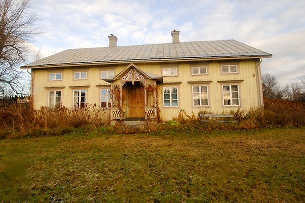 Rise 6:8. Villa i gul panel med beige detaljer, fönster med vita spröjs, förstukvist med snickarglädje, dubbeldörr i brunt.