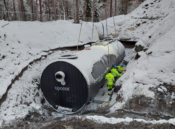 Bilden visar en stor rörformad tank som ligger i en grävd grop. Bredvid står tre vuxna personer som inte når högre än halvvägs upp på tanken.