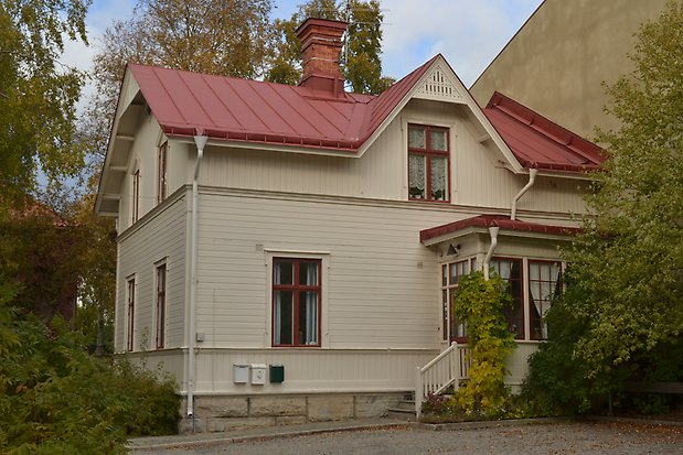 Villa i två våningar i ljusbeige liggande panel. Grund i sten. Röda fönster med spröjs. Punchveranda. Rött plåttak med skorsten i tegel.