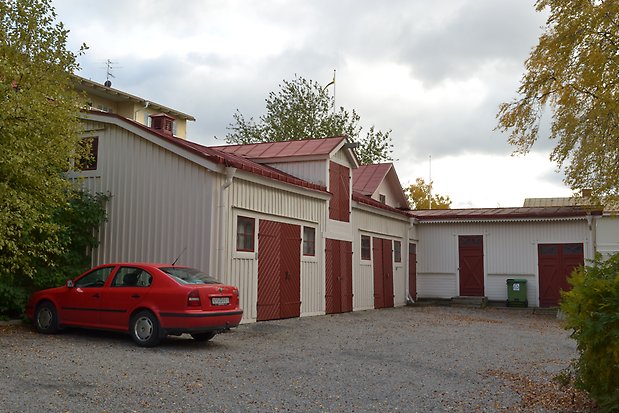 Uthus i L-vinkel i vit panel. Små, röda fönster med spröjs. Röda dubbelportar och enkelportar.