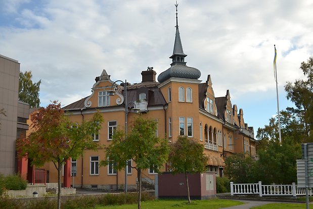 Stenbyggnad i tre våningar med tinnar och torn, gul puts, vita fönster med spröjs, svart plåttak.