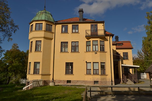 Stenhus i tre våningar i gul puts, tegeltak, bruna fönster med flera rutor och spröjs. Burspråk och ett torn med grönt plåttak. Stengrund.