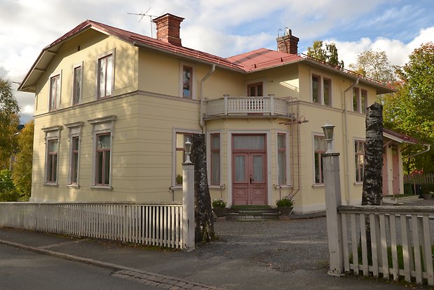 Villa i sten i ljusgul puts, vita detaljer och fönsterfoder, rosa fönster med spröjs. Rosa dubbeldörr med fönster. Balkong med vitt staket. Ljusrött plåttak med murade skorstenar.