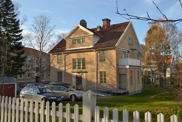 Villa i sten i tre våningar i beige puts. Gröna fönster med spröjs och många fönsterrutor. Rött tegeltak. Balkong med vitt staket. Brun dubbeldörr med fönster.