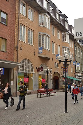 Tegelbyggnad i fyra våningar. Vita fönster med spröjs och många rutor. Burspråk på våningen två och tre. Svart plåttak med takkupor. Skyltfönster på nedervåningen.