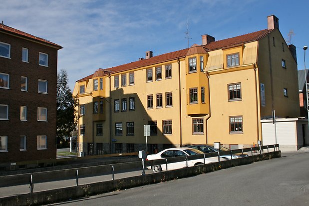 Byggnad i tre våningar i ljusgul puts. Två burspråk på våning två och tre i mörkgult. Rött tegeltak. Fönster och spröjs i brunt.